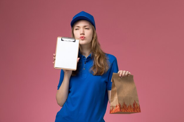 Front view female courier in blue uniform holding notepad and food package thinking on the light pink desk job worker service uniform company