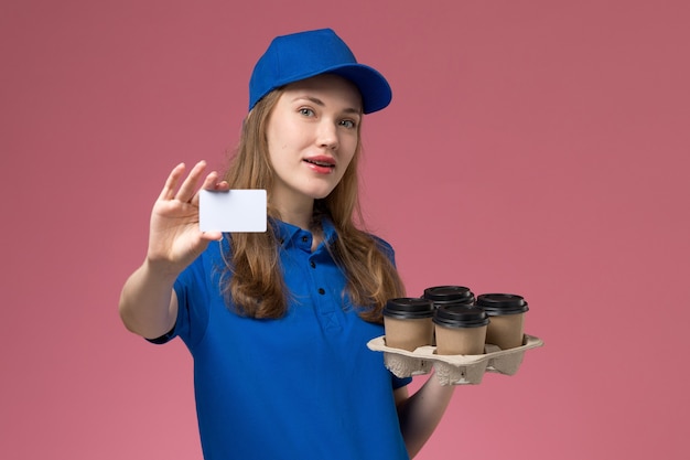 Front view female courier in blue uniform holding white card and brown delivery coffee cups on light-pink desk service uniform company