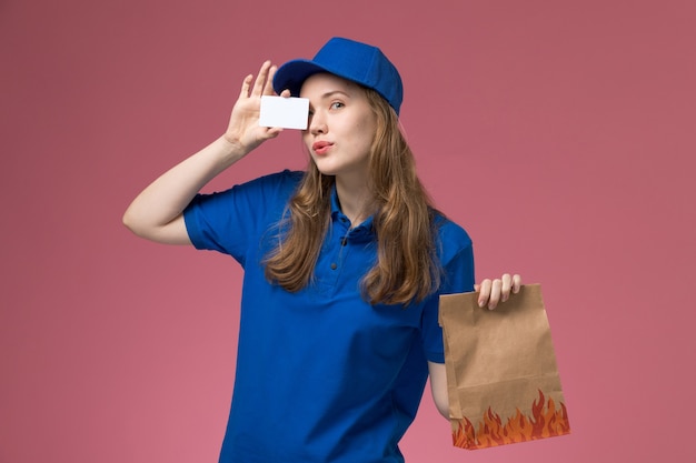 Front view female courier in blue uniform holding white card and food package on pink desk service uniform company job worker