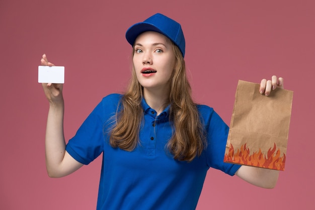 Front view female courier in blue uniform holding white card and food package with surprised expression on pink light desk service uniform company job