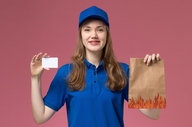 Вид спереди женщина-курьер в синей униформе улыбается, держит белую карточку и пакет с едой на розовом светлом столе, служба униформы, работа компании
