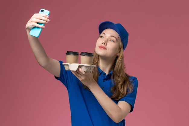 Front view female courier in blue uniform taking a selfie with brown delivery coffee cups on the pink desk service uniform company worker