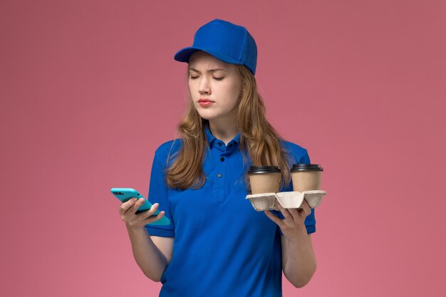 Вид спереди женщина-курьер в синей форме, использующая свой телефон с коричневыми кофейными чашками для доставки на розовом столе, служебная форма, работа компании