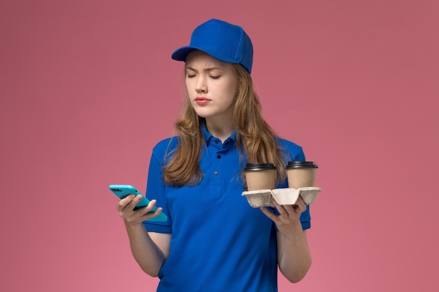 ピンクのデスクサービスの仕事の制服会社の仕事で茶色の配達コーヒーカップを保持している彼女の電話を使用して青い制服を着た女性の宅配便の正面図