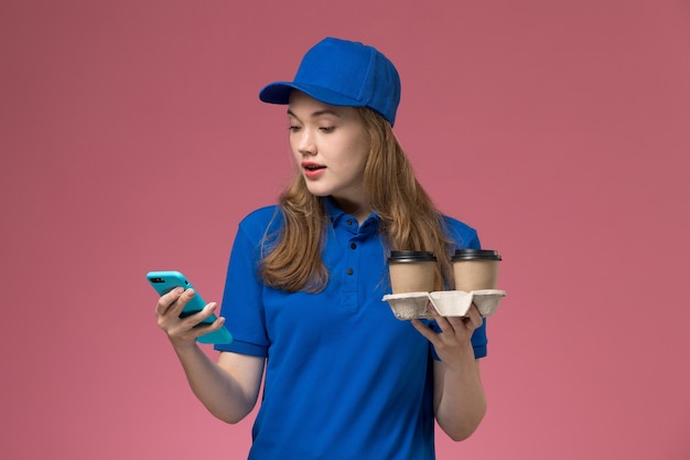 Front view female courier in blue uniform using her phone holding brown delivery coffee cups on pink desk service job uniform company