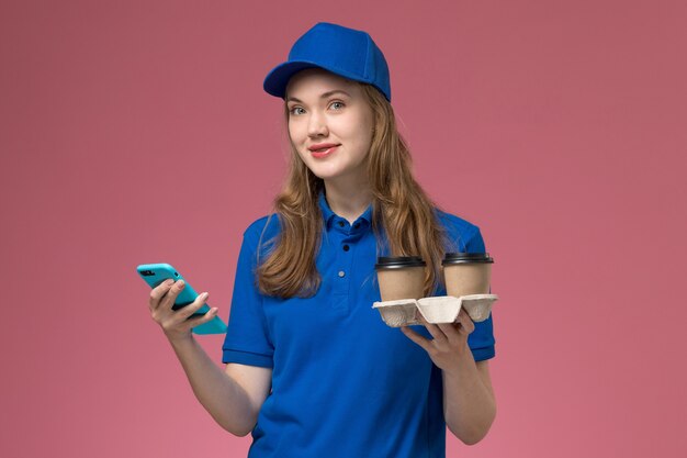 Front view female courier in blue uniform using her phone with smile holding brown delivery coffee cups on pink desk service job uniform company job