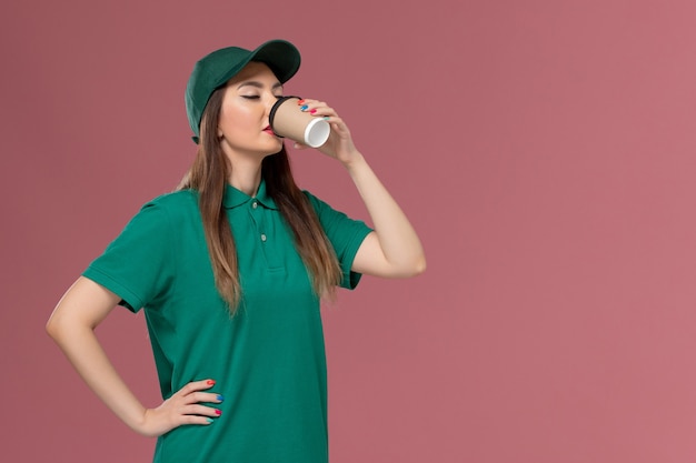 Front view female courier in green uniform and cape drinking coffee on the light-pink wall service job uniform delivery
