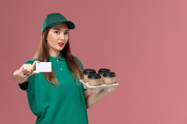front view female courier in green uniform and cape holding card and delivery coffee cups on pink desk service uniform delivery