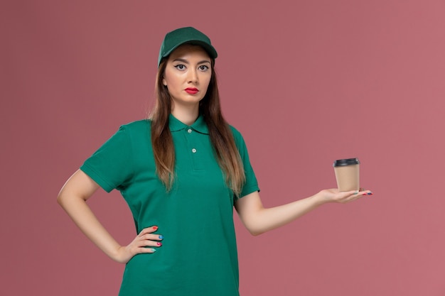 Вид спереди женщина-курьер в зеленой униформе и плаще, держащая чашку кофе для доставки на светло-розовой стене, служба доставки униформы