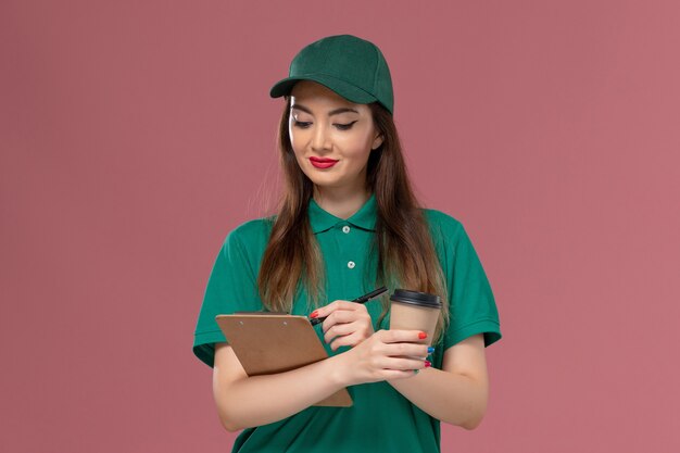 Front view female courier in green uniform and cape holding delivery coffee cup and notepad pen on light-pink wall company service job uniform delivery