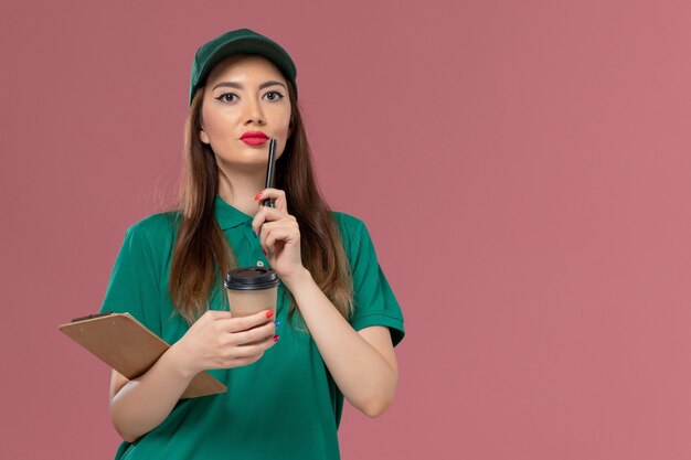 Front view female courier in green uniform and cape holding delivery coffee cup and notepad pen on pink wall company service job uniform delivery