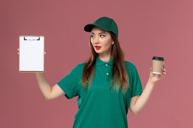 Front view female courier in green uniform and cape holding delivery coffee cup and notepad on pink desk service job uniform delivery work worker female company