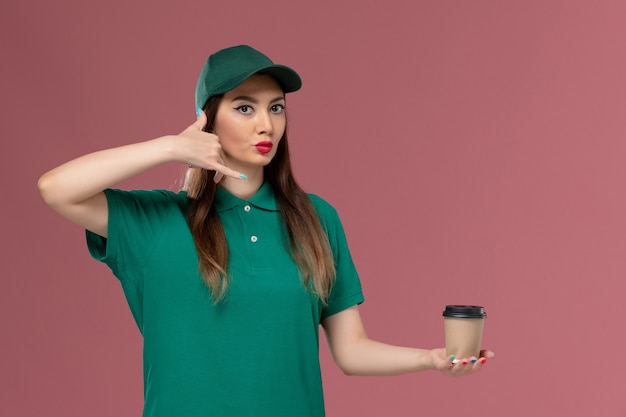 Free photo front view female courier in green uniform and cape holding delivery coffee cup on pink wall service job uniform delivery female work