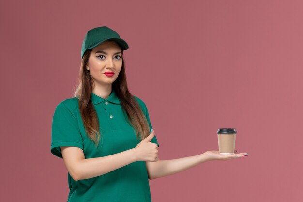 Front view female courier in green uniform and cape holding delivery coffee cup on pink wall service job uniform delivery worker female