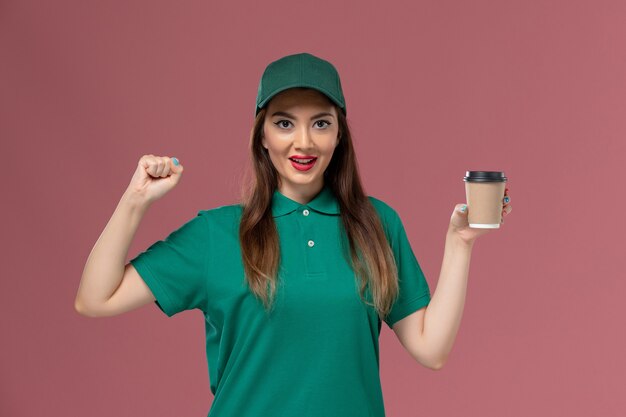 Front view female courier in green uniform and cape holding delivery coffee cup on pink wall service job uniform delivery worker