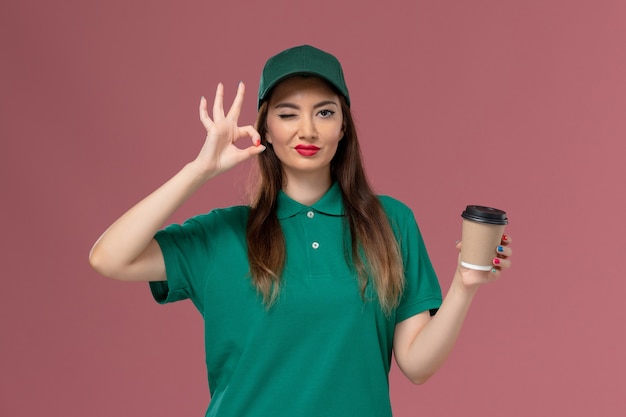 녹색 유니폼과 케이프 핑크 벽 서비스 유니폼 배달 작업자 작업에 배달 커피 컵을 들고 전면보기 여성 택배