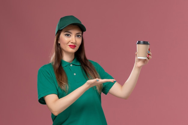 Front view female courier in green uniform and cape holding delivery coffee cup smiling on pink wall service job uniform delivery work