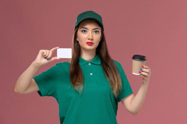 Front view female courier in green uniform and cape holding delivery coffee cup with card on pink wall service job uniform delivery work