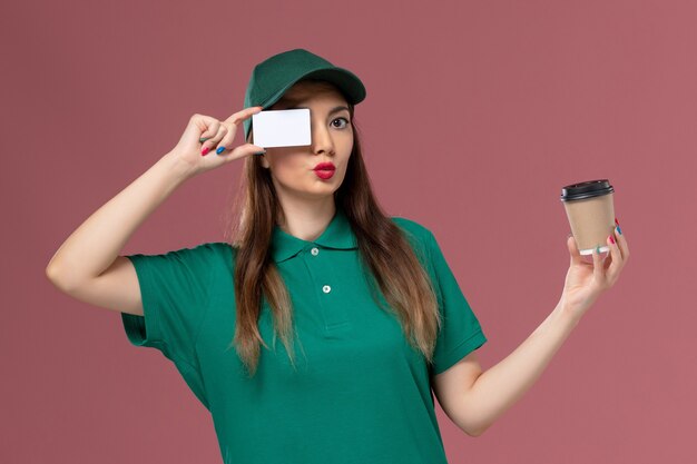 Front view female courier in green uniform and cape holding delivery coffee cup with card on the pink wall service job uniform delivery work