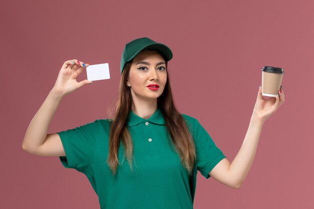 緑のユニフォームとピンクの壁のサービスジョブユニフォーム配達のカードと配達コーヒーカップを保持している岬の正面図の女性の宅配便