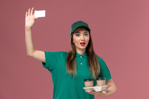 녹색 유니폼과 케이프 핑크 벽 서비스 작업 유니폼 배달 아가씨 배달 커피 컵과 카드를 들고 전면보기 여성 택배