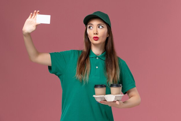 Front view female courier in green uniform and cape holding delivery coffee cups and card on pink wall service job uniform delivery worker