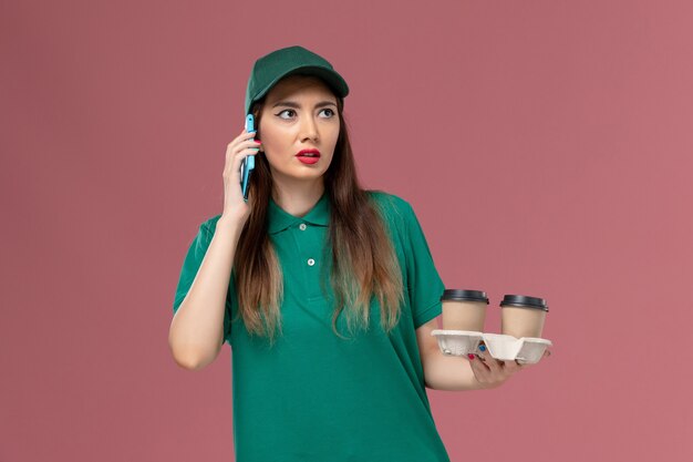 Вид спереди женщина-курьер в зеленой форме и накидке с доставкой кофейных чашек и ее телефоном на розовом столе.