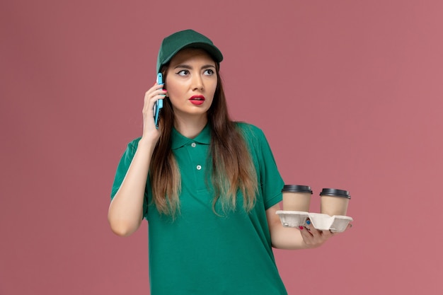 Front view female courier in green uniform and cape holding delivery coffee cups and her phone on pink desk service uniform delivery