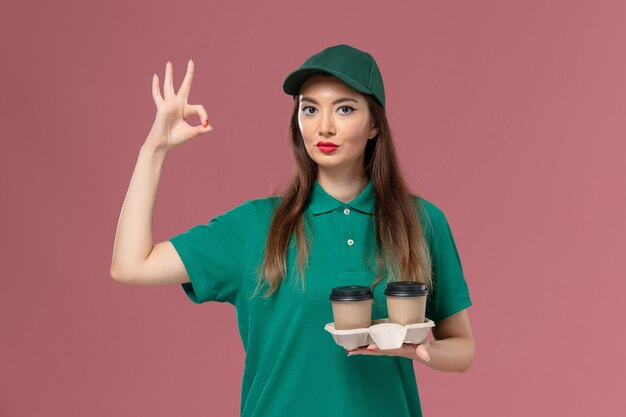 Вид спереди женщина-курьер в зеленой униформе и накидке с доставкой кофейных чашек на светло-розовом столе.