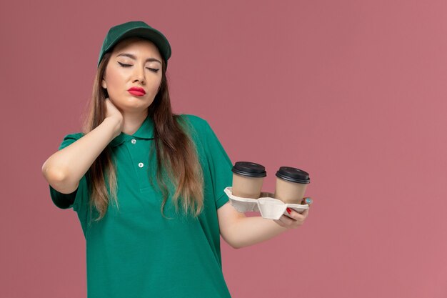 Вид спереди женщина-курьер в зеленой униформе и накидке с доставкой кофейных чашек на светло-розовой стене Доставка униформы компании