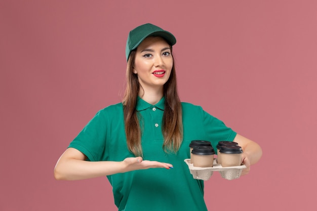 Front view female courier in green uniform and cape holding delivery coffee cups on the light pink wall service uniform delivery job