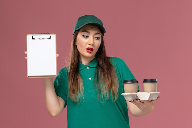 Front view female courier in green uniform and cape holding delivery coffee cups and notepad on light-pink desk service job uniform delivery