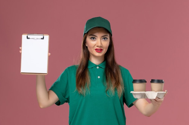 Corriere femminile di vista frontale in uniforme verde e mantello che tiene le tazze di caffè di consegna e blocchetto per appunti sull'operaio di consegna dell'uniforme di lavoro di servizio della parete rosa