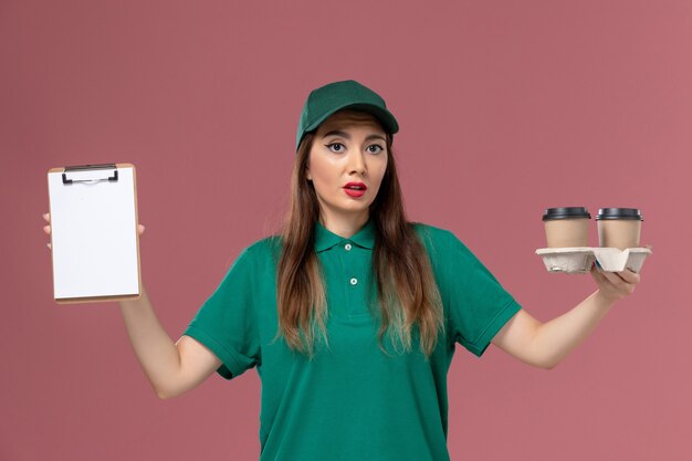 Front view female courier in green uniform and cape holding delivery coffee cups and notepad on pink wall service job uniform work delivery