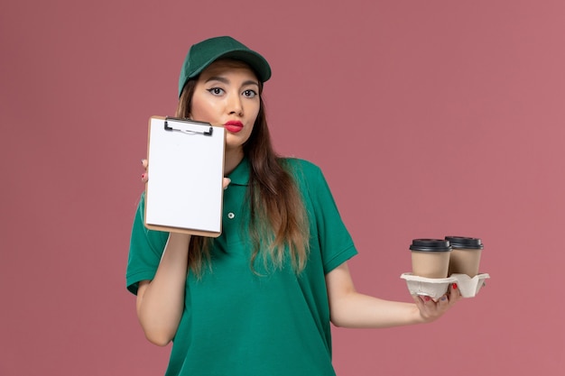 緑の制服を着た正面図の女性の宅配便とピンクの壁のサービスの仕事の仕事の均一な配達の配達コーヒーカップとメモ帳を保持している岬