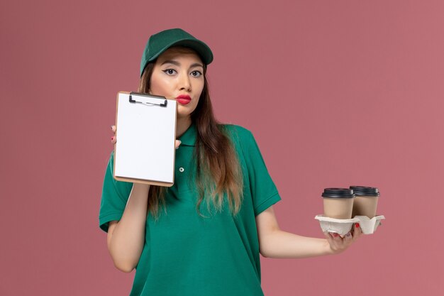 Front view female courier in green uniform and cape holding delivery coffee cups and notepad on pink wall service job work uniform delivery