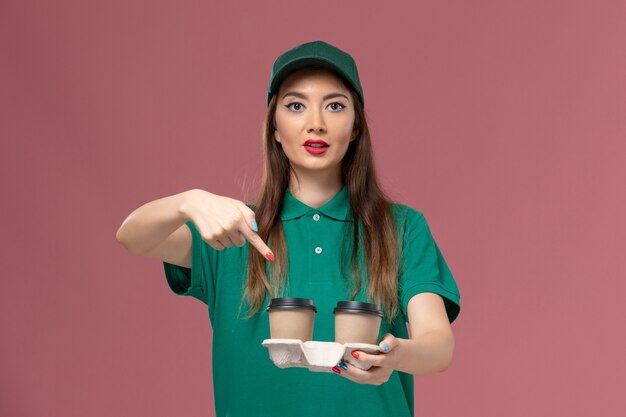 Front view female courier in green uniform and cape holding delivery coffee cups on pink wall company service job uniform delivery female