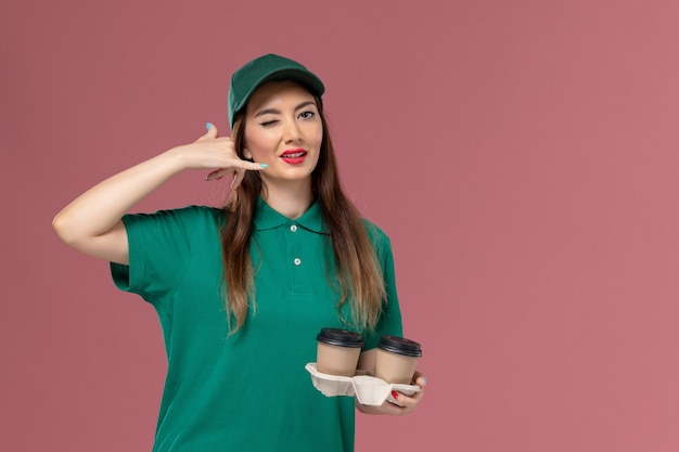 Front view female courier in green uniform and cape holding delivery coffee cups on pink wall company service job uniform delivery worker female work