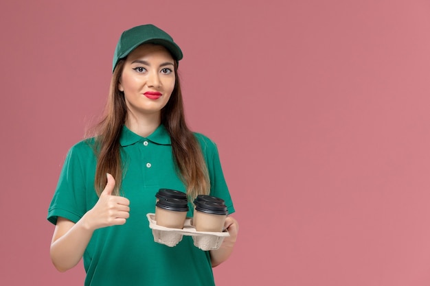 Free photo front view female courier in green uniform and cape holding delivery coffee cups on pink wall service company uniform delivery job