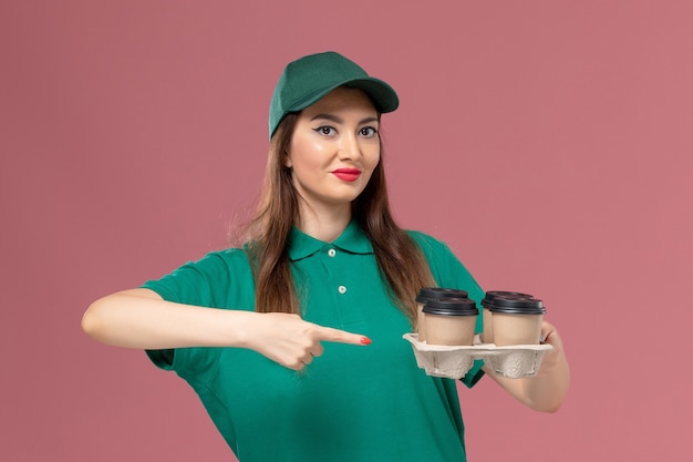 front view female courier in green uniform and cape holding delivery coffee cups on pink wall service uniform delivery job lady