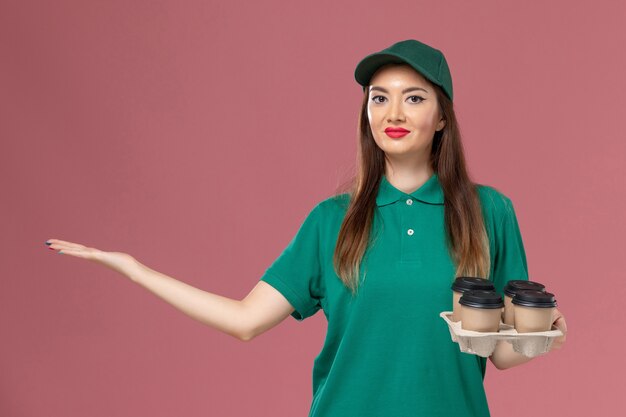 front view female courier in green uniform and cape holding delivery coffee cups on the pink wall service uniform delivery job work