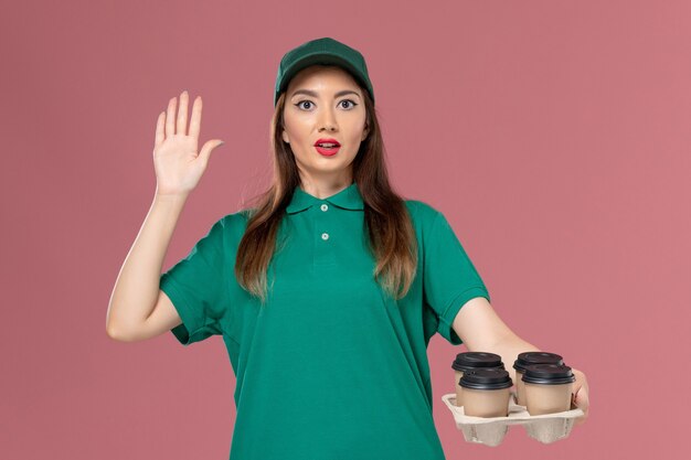 Вид спереди женщина-курьер в зеленой униформе и плаще, держащая кофейные чашки доставки на розовой стене, работа по доставке формы работника службы