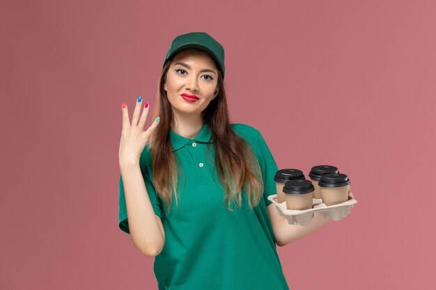 Вид спереди женщина-курьер в зеленой униформе и накидке с доставкой кофейных чашек, слегка улыбаясь на розовой стене.