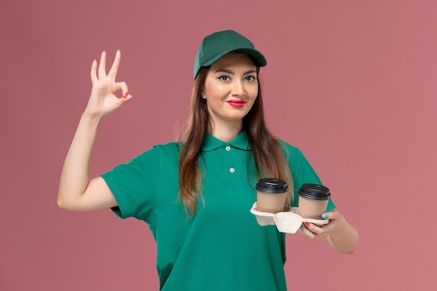 녹색 유니폼과 케이프 핑크 벽 회사 서비스 작업 유니폼 배달에 미소 배달 커피 컵을 들고 전면보기 여성 택배