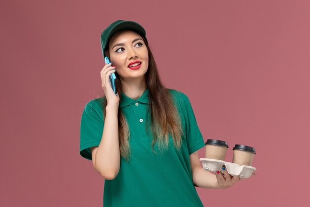 Front view female courier in green uniform and cape holding delivery coffee cups talking on the phone on pink desk service uniform delivery