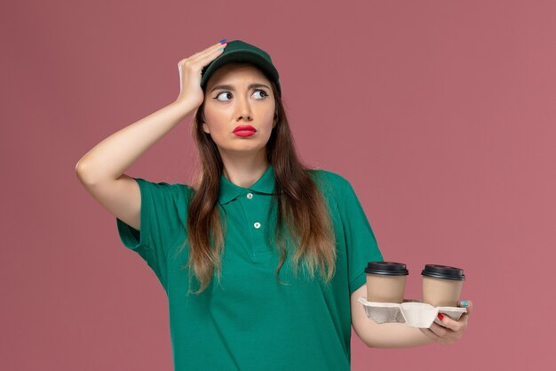 Вид спереди женщина-курьер в зеленой униформе и накидке держит доставку кофейных чашек, думая о светло-розовой стене, служба доставки униформы компании