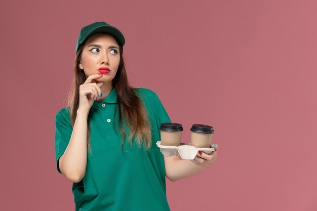 Front view female courier in green uniform and cape holding delivery coffee cups thinking on pink wall company service job uniform delivery worker