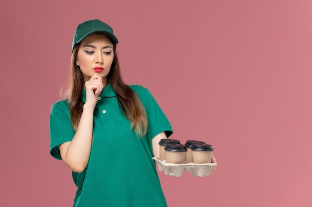 Front view female courier in green uniform and cape holding delivery coffee cups thinking on pink wall service uniform delivery job