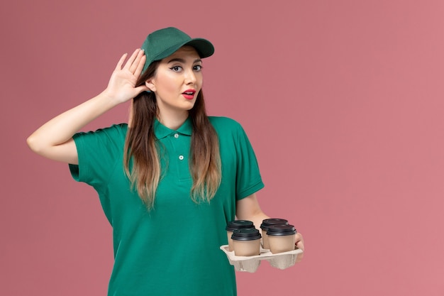 Вид спереди женщина-курьер в зеленой форме и накидке с доставкой кофейных чашек пытается услышать на розовой стене служба доставки униформы