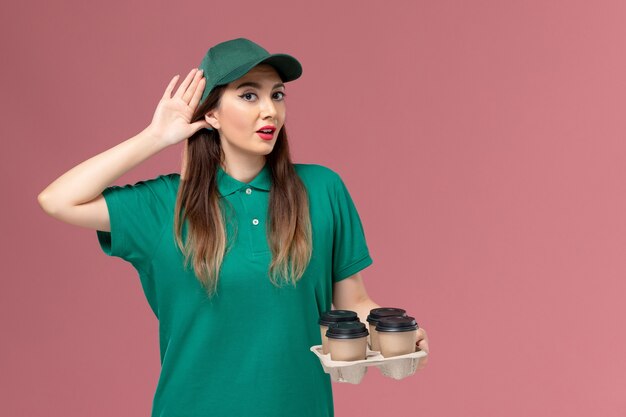 Front view female courier in green uniform and cape holding delivery coffee cups tryign to hear on pink wall service uniform delivery job